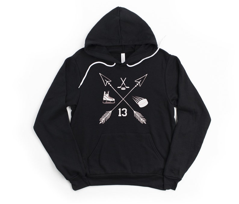 May include: Black hooded sweatshirt with a white graphic featuring crossed arrows, a hockey skate, a hockey stick and puck, and the number 13. The sweatshirt has a front pocket and white drawstring.