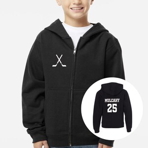 May include: A black zip-up hoodie featuring a white graphic of two crossed hockey sticks on the left chest. An inset circle shows the back of the hoodie, which displays the name MULCAHY in white block letters above the number 25 in white.