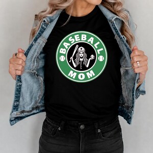 May include: Black t-shirt with a green and white graphic that says "Baseball Mom". The graphic features a baseball bat, a baseball, and a woman wearing a baseball cap. The shirt is being worn with a denim jacket.