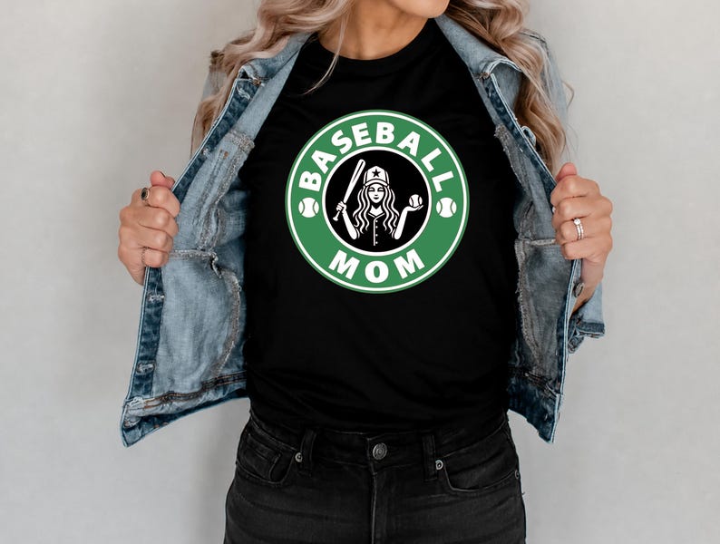 May include: Black t-shirt with a green and white graphic that says "Baseball Mom". The graphic features a baseball bat, a baseball, and a woman wearing a baseball cap. The shirt is being worn with a denim jacket.