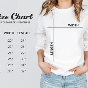 K&ouml;nnte beinhalten: Ein wei&szlig;es Unisex-Crewneck-Sweatshirt mit einer Gr&ouml;&szlig;entabelle. Die Tabelle listet die Gr&ouml;&szlig;en S bis 3XL mit den entsprechenden Breiten- und L&auml;ngenma&szlig;en in Zoll auf. Das Model tr&auml;gt das Sweatshirt und eine blaue Jeans.
