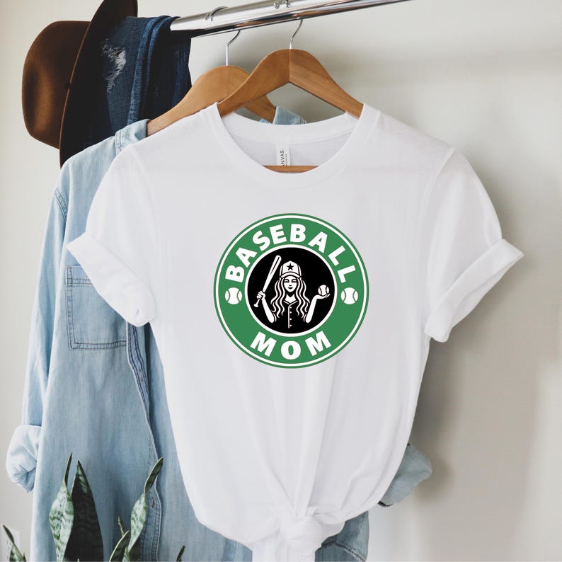 May include: White t-shirt with a green and black graphic. The graphic features a woman holding a baseball bat and ball, surrounded by the words "BASEBALL MOM" in a circular design. The shirt is hanging on a wooden hanger.