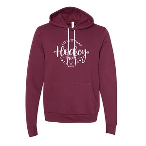 K&ouml;nnte beinhalten: Ein weinroter Hoodie mit Fronttasche und Kapuze mit Kordelzug. Der wei&szlig;e Text auf der Vorderseite lautet "Livin' that Hockey Mom Life" mit Hockeyschl&auml;gern und Sternen. Ein bequemes Sweatshirt f&uuml;r Hockeyfans.