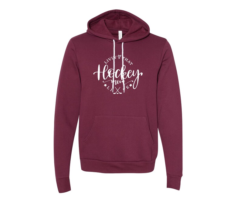 K&ouml;nnte beinhalten: Ein weinroter Hoodie mit Fronttasche und Kapuze mit Kordelzug. Der wei&szlig;e Text auf der Vorderseite lautet "Livin' that Hockey Mom Life" mit Hockeyschl&auml;gern und Sternen. Ein bequemes Sweatshirt f&uuml;r Hockeyfans.