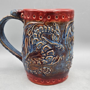 Op de afbeelding: Een handgemaakte keramische mok met een drakenontwerp. De mok heeft een blauw en bruin drakenreliëf tegen een donkerbruine achtergrond. De rand en de basis zijn dieprood. De handgreep is blauw.