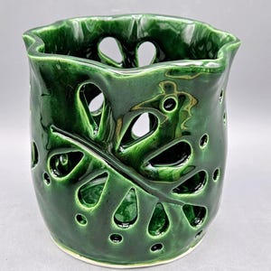 Könnte beinhalten: Eine glänzende, smaragdgrüne Keramikvase mit einem einzigartigen, blattartigen Ausschnitt-Design. Die Vase hat einen wellenförmigen Rand und eine glatte, reflektierende Oberfläche. Das dekorative Stück eignet sich für Blumen oder als eigenständiges Kunstobjekt.