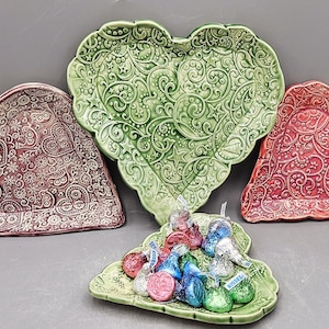 May include: Four heart-shaped ceramic dishes in shades of green, red, and burgundy, with embossed floral and star patterns. A small green leaf-shaped dish holds wrapped sweets. Decorative serving pieces.