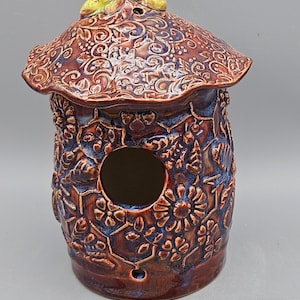 May include: A ceramic birdhouse with a detailed design of flowers, leaves, and bees. The birdhouse is painted in shades of brown and blue with a green roof.