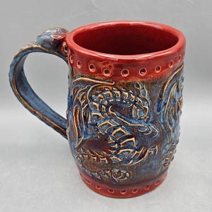 May include: A handcrafted ceramic mug with a dragon design. The mug features a red interior and rim, with a textured blue and brown exterior. The handle is blue and has a decorative element. The mug is approximately 5 inches tall.