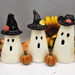 May include: Three white ceramic ghost figurines with hollow eyes and mouths. Each ghost wears a black hat; two are pointed witch hats with small red or yellow flowers, and one is a black cowboy hat with a small red flower on the brim.