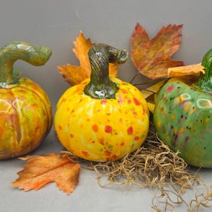 May include: Three ceramic pumpkins with glossy finishes. The left pumpkin is orange and yellow with a green stem. The center pumpkin is bright yellow with orange and green speckles and a dark green stem. The right pumpkin is green with pink and yellow spots.