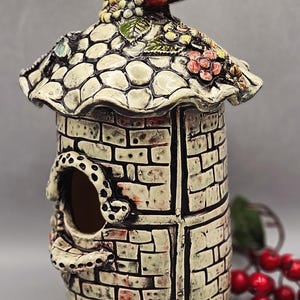 May include: A ceramic birdhouse featuring a red bird on the roof. The birdhouse has a cylindrical shape with a brick design and a decorative roof with floral details. The birdhouse has a circular opening.