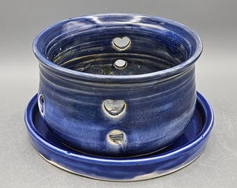 May include: A handmade, dark blue ceramic garlic keeper with a matching saucer. The keeper has three heart-shaped cutouts and a textured surface. The ceramic piece is glazed with a glossy finish.