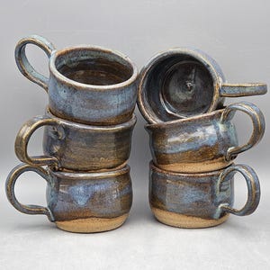May include: A stack of six handmade ceramic mugs. The mugs have a blue and brown glaze with a rustic, textured appearance. Each mug has a handle and a rounded base. The mugs are stacked on top of each other.