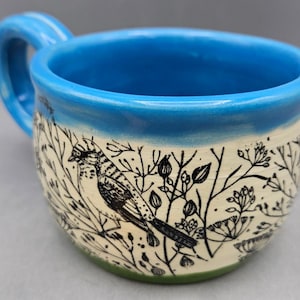 May include: A ceramic mug with a bright blue glazed rim and handle. The body of the mug is cream-colored and features a detailed black ink illustration of a bird perched among various wild plants, flowers, and stems wrapping around the exterior.