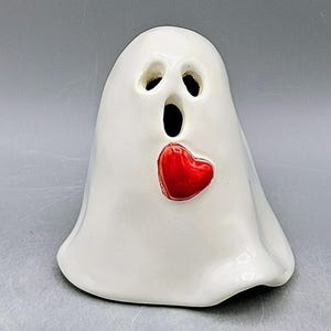 May include: A white ceramic ghost figurine with a red heart. The ghost has three black eye holes and a large open mouth. The red heart is positioned on the ghost's chest. The figurine is a Halloween or Valentine's Day decoration.