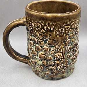 May include: A ceramic mug with a brown glazed finish and a textured exterior featuring a repeating pattern of small, raised skull shapes. The mug has a curved handle on the left side and a smooth, cylindrical rim at the top, showcasing a glossy surface.