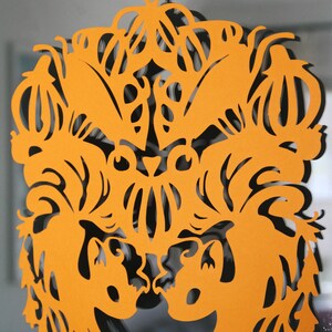 Könnte beinhalten: Orangefarbene Papier-Ausschnittdekoration mit einem symmetrischen Design. Das Kunstwerk zeigt stilisierte Tier- oder Blumenformen, mit dem Wort "@AKTHAUSEN" in der oberen linken Ecke. Die Dekoration wird von einer blauen Schnur gehalten.