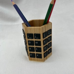 May include: A hexagonal wooden pencil holder with a unique design. The holder is made of wood and has black keyboard keys attached to the sides. Two pencils, one purple and one green and red, are in the holder.