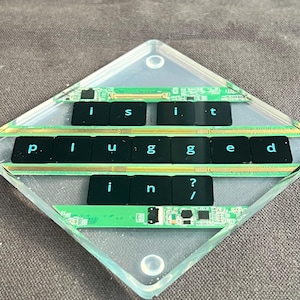 May include: A diamond-shaped clear resin coaster embedded with green circuit board strips and black computer keyboard keys. The keys are arranged to spell out the phrase "is it plugged in?" in three horizontal rows, set against the clear material.