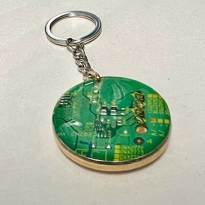 Puede incluir: Un llavero redondo con un diseño de placa de circuito verde encapsulado en resina transparente. El llavero tiene un borde de metal dorado y un llavero y cadena plateados. La placa de circuito tiene detalles amarillos y blancos.