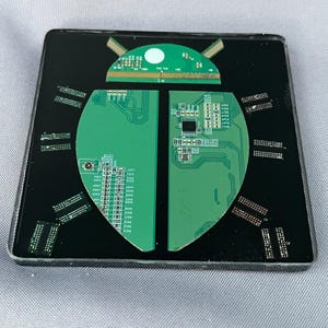 May include: A square black coaster featuring a green printed circuit board shaped like a beetle. The design includes a semi-circular head piece and two wing-like body sections with visible electronic components, traces, and small metallic pins.