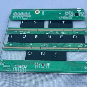May include: A rectangular clear acrylic coaster featuring embedded green circuit boards and black computer keyboard keys. The keys are arranged to spell out the text "I S I T T U R N E D O N ?" across three horizontal rows separated by the circuit boards.