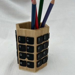 May include: A wooden pencil holder with a hexagonal shape. The holder is made of wood and has a design of computer keyboard keys. There are four pencils in the holder: one red, one blue, one green, and one purple.