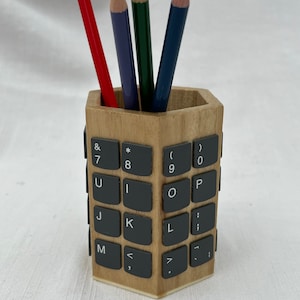 May include: Wooden pencil holder shaped like a keyboard with black keys. The holder has four pencils inside: red, purple, green, and blue.
