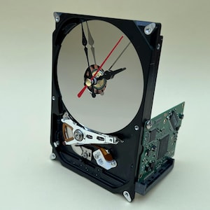 May include: A distinctive clock crafted from a recycled hard drive. The clock face is the drive's reflective surface, with black hands and a red second hand. The clock has a black frame, and a green circuit board is attached.