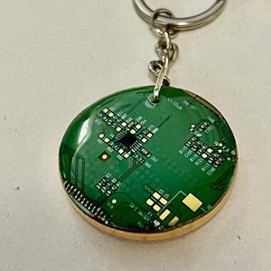 Puede incluir: Un colgante de llavero redondo con un diseño de placa de circuito verde y detalles dorados. El colgante está encapsulado en resina transparente y tiene un borde de madera de color dorado. El llavero tiene un anillo de metal plateado.