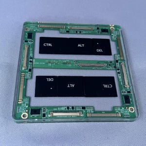 May include: A rectangular green printed circuit board frame featuring multiple metallic connectors and integrated components. Two black rectangular panels are embedded within the frame, each displaying the white text CTRL, ALT, and DEL.
