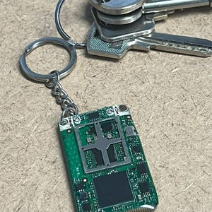 以下が含まれることがあります： 緑色の基板キーホルダーとシルバーの金具。キーホルダーは一連の銀色の鍵に付いています。基板にはさまざまな電子部品と長方形の黒い部品があります。