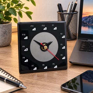 May include: A square black analog clock with a circular gray center and a red second hand. The clock face features white function key labels from F1 to F12 arranged in a circle, each accompanied by a small white icon representing computer commands.