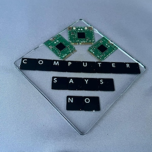 May include: A clear diamond-shaped coaster featuring three green computer circuit boards arranged at the top and black keyboard keys spelling out the phrase COMPUTER SAYS NO in three rows across the center of the transparent surface.