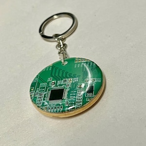 May include: A green circuit board keyring with a silver ring. The keyring has a rounded, oval shape with a wooden border. The circuit board design features intricate lines and components.