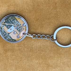 May include: A silver keyring with a round, gold-coloured charm featuring a yin yang symbol on a circuit board design. The text "FU5856  LE1394" is visible on the charm.