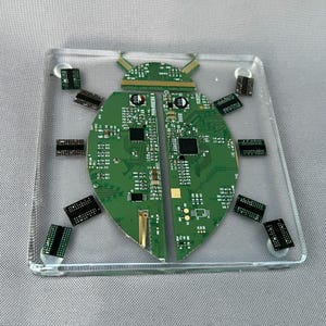 May include: A square clear resin coaster featuring an embedded insect shape constructed from green printed circuit boards. The design includes two large semi-circular body segments and smaller rectangular electronic components as legs.