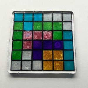 May include: Square coaster with a grid of colourful squares in shades of blue, green, pink, purple, silver, and gold. The squares are encased in a clear, square border.