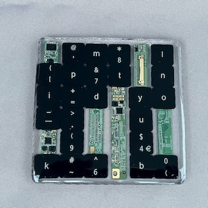 May include: A square coaster made of clear resin containing a collection of black computer keyboard keys and green electronic circuit board components. The keys feature white printed characters including letters, numbers, and symbols arranged in a grid.