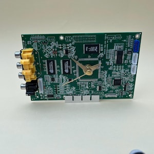 Può includere: Un orologio unico realizzato con un circuito stampato verde. L'orologio presenta lancette dorate e un quadrante dorato. Il circuito stampato ha vari componenti elettronici e connettori. L'orologio è una miscela creativa di tecnologia e misurazione del tempo.