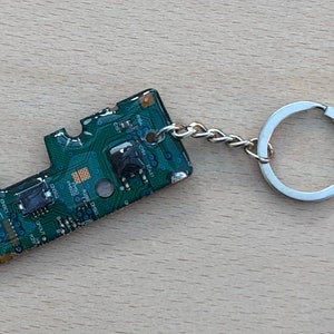 Peut inclure: Un porte-clés en forme de circuit imprimé vert avec un anneau de clé argenté. Le circuit imprimé comporte divers composants et du texte, notamment "C501", "C502" et "R200".