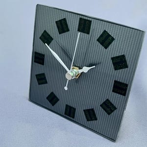 May include: A square wall clock with a dark gray, vertically striped glass face. The clock features twelve black square markers, each containing raised dot patterns, arranged in a circular formation. It has three thin white hands at the center.
