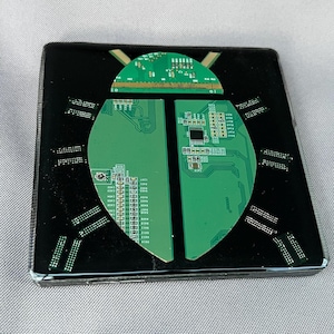May include: A square coaster featuring a beetle design crafted from green printed circuit board fragments embedded in a glossy black resin base. The insect shape is composed of a semi-circular head and two wing-like body sections with small rectangular circuit pieces forming the legs.