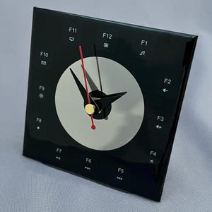 May include: A square black wall clock with a circular silver center. The clock face features white function key labels from F1 to F12, each accompanied by a small icon. It has black hour and minute hands and a thin red second hand with a gold center.