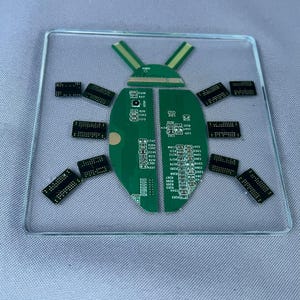 May include: A square clear glass coaster featuring a decorative insect shape assembled from green printed circuit board fragments. The design includes two green body halves, a head piece, and eight small black rectangular electronic components.