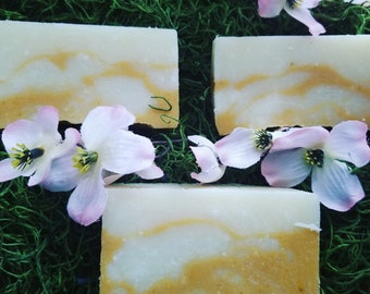 May include: Three bars of handmade soap with a white base and swirls of yellow. The soap is displayed on a bed of green moss with pink flowers.
