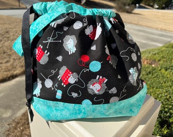May include: A drawstring project bag featuring a black fabric body with a pattern of white sheep wearing red sweaters, blue yarn balls, and knitting needles. The bag has a turquoise marbled fabric base and a matching turquoise drawstring top.