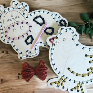 May include: Two wooden animal shapes with holes for stringing yarn. The shapes are a cow and a bird. The cow is white with black spots and the bird is white with yellow accents. A pink yarn needle is included.