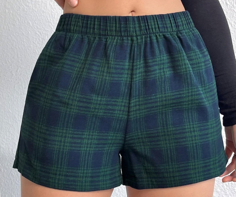 K&ouml;nnte beinhalten: Gr&uuml;n-marine karierte Shorts mit elastischem Bund. Die Shorts haben ein klassisches Karomuster in Gr&uuml;n- und Marineblaut&ouml;nen. Der Stoff scheint ein weiches, gewebtes Material zu sein.